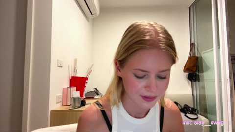 Media: A video of a blonde woman with fair skin and straight hair in a white tank top, standing in a modern, minimalist bathroom with a mirror, light fixtures, and a red organizer.