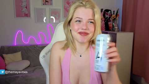 Media: A video of a young blonde woman with fair skin, smiling, wearing a pink halter top, holding a can, in a cozy, pink-lit room with decor and a white chair.