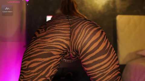 Video of a woman with light skin and long brown hair, wearing a tight, zebra-patterned bodysuit that reveals her buttocks and vagina. The background is dimly lit with a blurred, warm-colored wall.