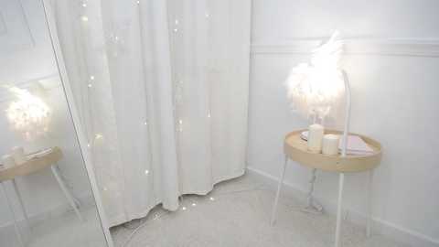 A video of a minimalist, white-walled room with a large mirror, a wooden side table, and a white feather lamp casting soft light.