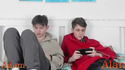 Video of two teenage boys, one in a beige hoodie, the other in a red blanket, playing a handheld video game on a bed, white headboard, blue artwork on the wall.