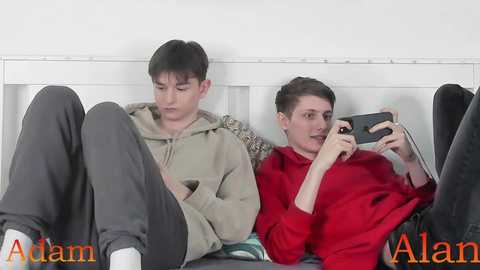 Video of two young men, one in a beige hoodie, sitting side by side on a bed, looking at a phone. Text: \"Adam Alar.\