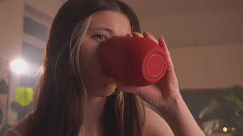 Video of a young woman with long brown hair, wearing a red beanie, drinking from a red cup. The background is blurred, featuring a dimly lit room with a window and potted plants.
