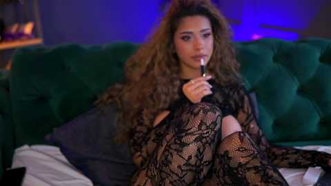 Media: Video of a young woman with long, wavy brown hair, wearing a black lace bodysuit and thigh-high stockings, smoking a cigarette, sitting on a green velvet bed in a dimly lit room.