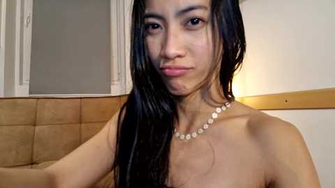 Video of an Asian woman with long, wet black hair, wearing a pearl necklace, smiling, standing in a beige-tiled bathroom with a tan couch.