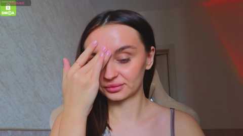 A video of a young woman with straight, dark hair, wearing a gray spaghetti-strap top, touching her forehead with her right hand, indoors with a textured gray wall and a doorway in the background.
