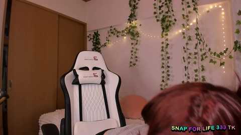 Media: Video of a white gaming chair with a cushioned headrest in a cozy, dimly lit room adorned with fairy lights and green vines. A person with light brown hair is partially visible in the foreground.