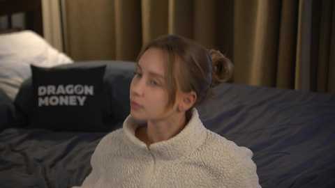 Media: Video of a young woman with light skin and brown hair in a bun, wrapped in a white blanket, sitting on a dark bed. A black pillow with \"DRAGON MONEY\" text is visible.