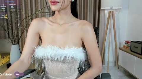 Media: Video of an Asian woman with fair skin, dark hair, wearing a strapless, feather-trimmed, sheer white dress, seated indoors next to a modern white lamp and a wooden stool.