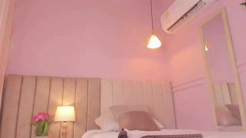 Video of a minimalist, pastel pink bedroom with a beige tufted headboard, white bed, and two pendant lamps.