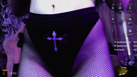 Video of a person wearing black fishnet stockings and a black thong with a white cross, displaying a pale, smooth abdomen. Background features a dark, dimly lit room with purple lighting and a digital clock.