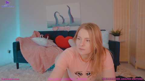 Media: A video of a young, fair-skinned woman with long, straight blonde hair, wearing a pink T-shirt, sitting on a plush white carpet in a brightly lit room with a dark blue couch, red heart-shaped pillow, and abstract artwork on the wall.