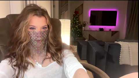 Video of a woman with long, wavy brown hair, wearing a leopard-print face mask, sitting on a brown couch in a modern living room with a TV, Christmas tree, and purple lighting.