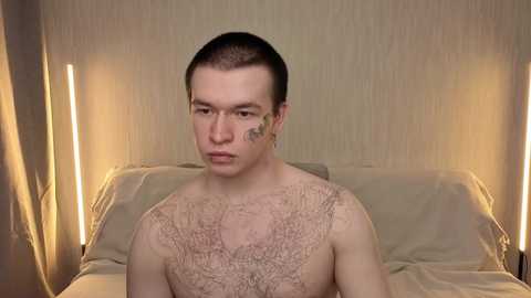 Media: Video of a young, fair-skinned, short-haired man with a small tattoo on his forehead, wearing a lace-up top, sitting on a beige bed with a soft, textured headboard, in a dimly lit, minimalist bedroom.