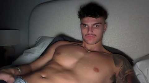 Video of a shirtless, muscular young man with short, curly hair and a stern expression, lying on a bed with white pillows and a beige headboard.