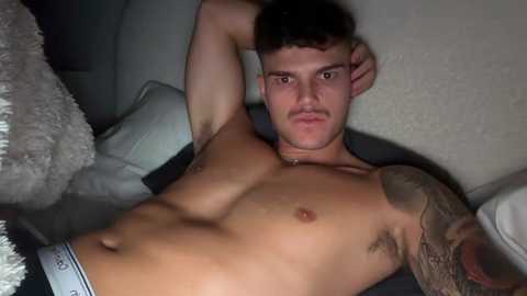 Video of a muscular, shirtless man with short dark hair and a tattoo on his right arm, lying on a bed with white pillows. He wears Calvin Klein underwear, with a neutral expression.