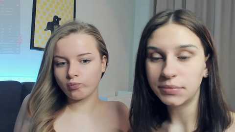 Video of two young women with fair skin and straight hair, one blonde, one brunette, both wearing minimal makeup, in a casual indoor setting with a yellow patterned wall and gray curtains.