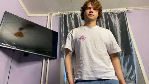 A video of a young white man with wavy brown hair, wearing a white t-shirt and blue jeans, standing in a room with a flat-screen TV on the left and a gray curtain behind him.