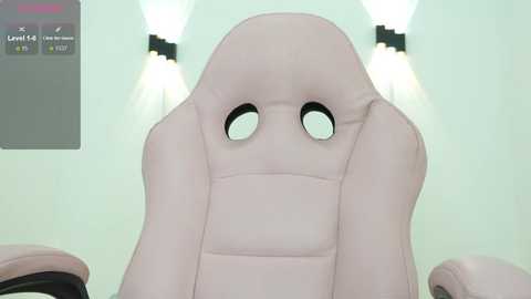 Video of a beige gaming chair with a cushioned backrest, featuring large, round, black eye-like cutouts, set against a minimalist white wall with two hanging lamps.