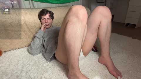 Video of a pale, short-haired man with a light gray sweater, reclining on a beige carpet, with his legs spread apart, revealing a glimpse of white underwear. Background shows a beige couch and green cloth.