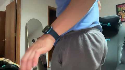 Media: Video of a person in a light blue shirt and gray sweatpants, playing video games in a dimly lit room with a gaming chair, wooden door, and framed art on the wall.