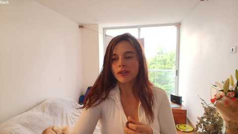 Media: Video of a young woman with long, straight brown hair, wearing a white shirt, standing in a bright, sunlit bedroom with white walls and a window.