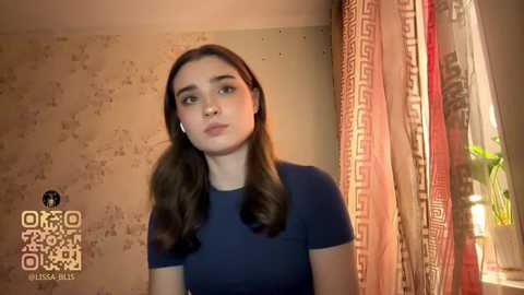 Video of a young woman with long brown hair in a dark blue top, standing in a room with patterned curtains and a plant, wearing white earphones.