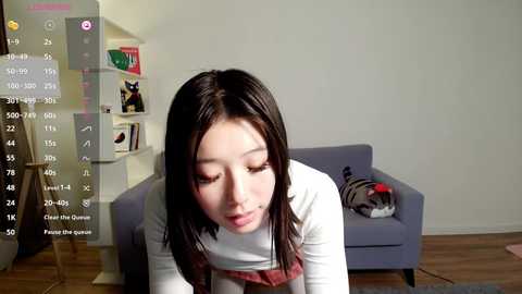 Video of a young Asian woman with long black hair, wearing a white turtleneck, leaning over a blue sofa in a minimalist living room.