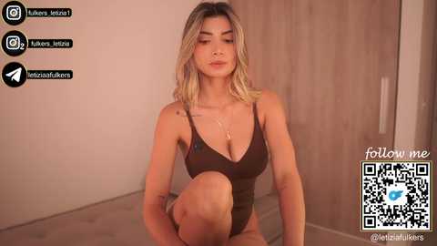 Media: Video of a blonde, light-skinned woman in a brown, deep V-neck bodysuit, sitting on a beige floor in a minimalist room. Social media icons and a QR code are visible.