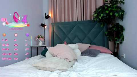 Video of a cozy, pastel-themed bedroom with a large bed adorned with white bedding, light pink pillows, and a plush teddy bear. The headboard is upholstered in teal, and there's a potted plant in the corner.