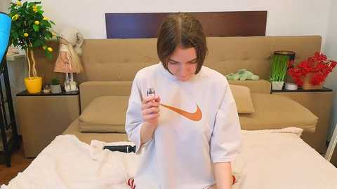Video of a woman with short brown hair, wearing a white sweatshirt with an orange Nike logo, smoking a cigarette on a beige bed with a white blanket, surrounded by potted plants and a wooden headboard in a cozy, well-lit room.