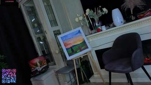 Video of an artist's studio with an easel holding a landscape painting, a white table cluttered with vases, and a dark grey chair.