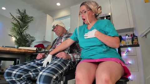 Media: Video of a middle-aged, white, bearded man in a plaid shirt, seated in a kitchen, wearing a white mask and gloves. Beside him, a blonde woman in a turquoise top and pink skirt, also wearing a mask and gloves, looks concerned.