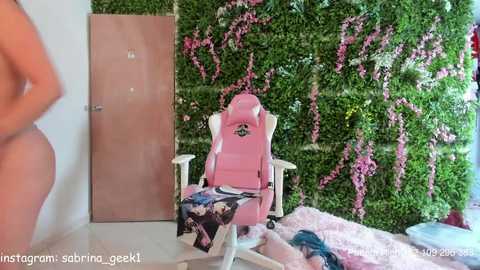 Video of a pink gaming chair with a floral blanket, surrounded by lush green foliage and pink flowers on a white wall, with a person's arm in the foreground.