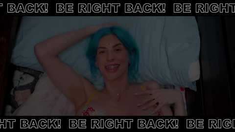 A video of a smiling woman with vibrant blue hair lying on a bed, wearing a floral-patterned top, with text overlaying the image saying \"I BE RIGHT BACK! BE RIGHT BACK! BE RIGHT BACK! BE RIGHT BACK!\