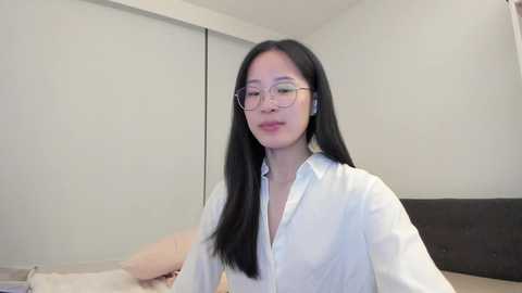 Video of an East Asian woman with long black hair, wearing glasses, a white button-up shirt, and a neutral expression, sitting on a beige bed in a minimalist, light-colored room with closed blinds.
