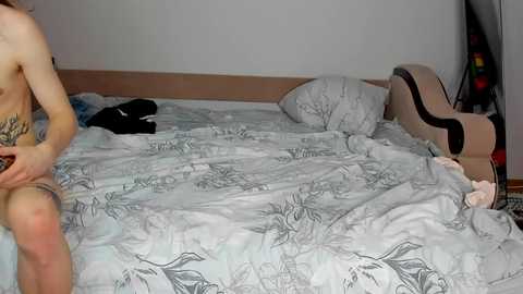 A video showing a nude woman with fair skin, tattooed legs, sitting on a bed with a light blue and white patterned duvet, in a cluttered, dimly-lit bedroom.