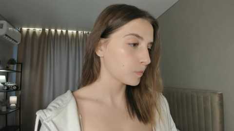 Video of a young Caucasian woman with fair skin, long brown hair, and a white shirt, standing indoors near a beige radiator, beige curtains, and a black shelf with books.