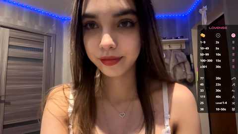 Video of a young woman with long brown hair, fair skin, and red lipstick, wearing a white lace bra, standing in a dimly lit bedroom with blue LED strip lights and a calendar on the right.