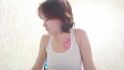 A video of a young East Asian woman with short, dark brown hair and a small pink rose tattoo on her shoulder. She's wearing a white tank top, looking downward. The background is a blurred, light-colored, textured wall.