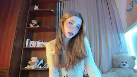 Video of a young woman with long, light brown hair, fair skin, and freckles, wearing a white off-shoulder top, leaning forward in a cozy room with wooden paneling, stuffed animals, and a teddy bear.