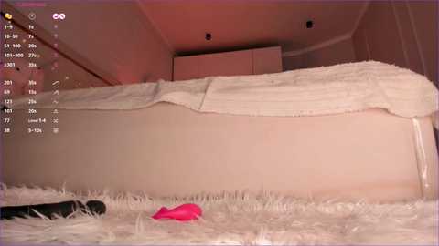 Video of a bedroom with a fluffy white rug, a bed with white sheets, and a pink vibrator on the floor. The room has beige walls and a ceiling with recessed lights.