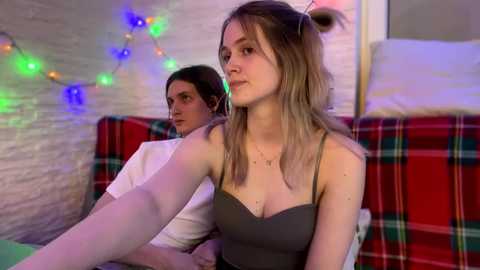 Video of a young woman with blonde hair in a gray top, sitting on a red plaid couch, next to a man with long dark hair in a white shirt, in a dimly lit room with colorful string lights.