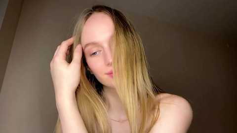 Video of a fair-skinned, slender woman with long, straight blonde hair partially covering her face, holding her hair with one hand. She has a delicate, almost serene expression. The background is a plain, light beige wall.