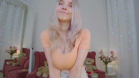 Video of a blonde woman with long, wavy hair, wearing a low-cut, peach-colored top, smiling in a cozy room with teddy bears, flowers, and soft lighting.