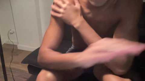 A video of a shirtless man with a muscular build, seated on a black chair, covering his face with his hands. He has light skin and is indoors, with a beige wall and a power outlet in the background.