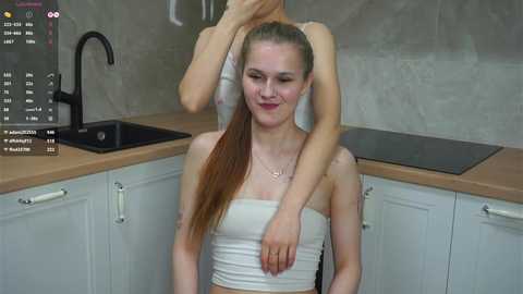 A video of a slender, fair-skinned young woman with long brown hair, wearing a white strapless top, being hugged by an unseen person in a modern kitchen with light wood countertops and white cabinets.