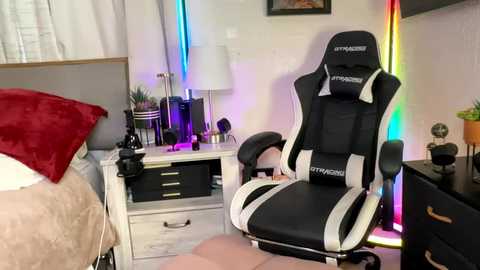 Video of a modern gaming room with a black and white gaming chair, a white desk, a red blanket, and colorful LED lights.