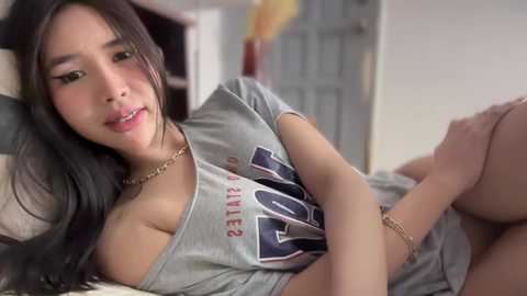 Video of an East Asian woman with long black hair, wearing a gray off-the-shoulder t-shirt, lounging on a bed in a casual, indoor setting.