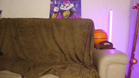 Video of a cozy living room featuring a beige couch draped with a brown blanket, a purple basketball, and a basketball-themed poster on the wall.